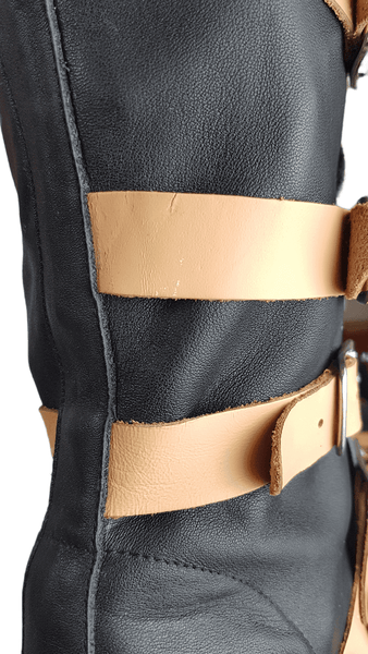 Side closeup view of a pair of black mid-calf leather boots wrapped with contrasting tan straps and antiqued metal buckles