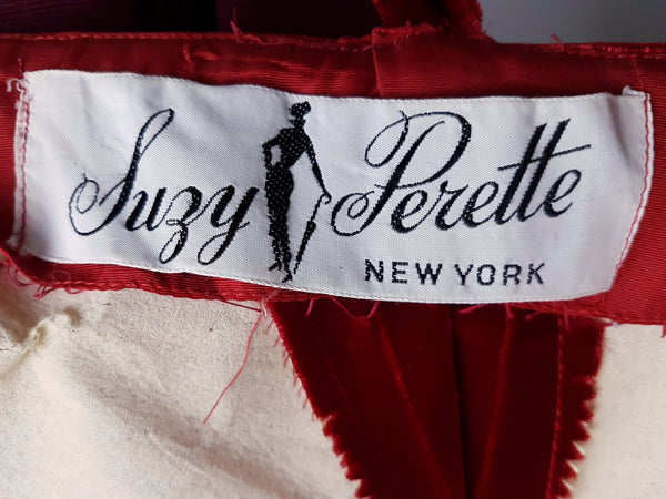 Label of Side bodice view of a circa mid-1950s Suzy Perette dark red velvet dress with square neckline, narrow straps and full skirt displayed on a mannequin 