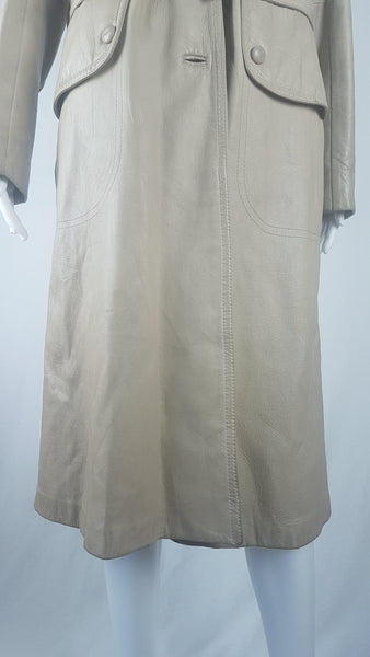 Front bottom hem view of a vintage 1970s sand-coloured leather trench coat with tonal buttons, shoulder tabs and a matching belt displayed on a mannequin
