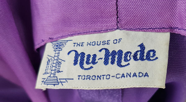 Label on a 1970s Nu-Mode purple jumpsuit with a draped hood, V-neckline, front zip and ties, long sleeves and wide legs displayed on a mannequin 