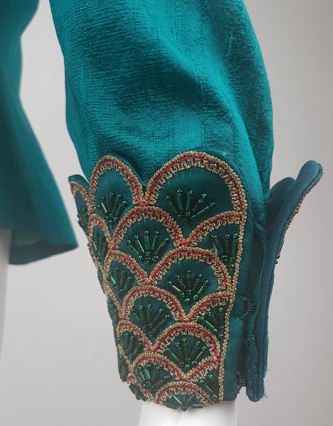Close-up of a deep teal silk cuff and cuff closure, decorated with overlapping scallops, metallic-gold braid, red cord and emerald beads.