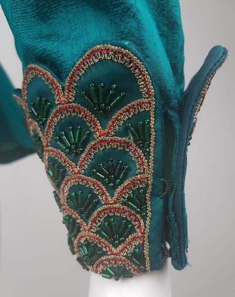 Close-up of a deep teal silk cuff and cuff closure, decorated with overlapping scallops, metallic-gold braid, red cord and emerald beads.