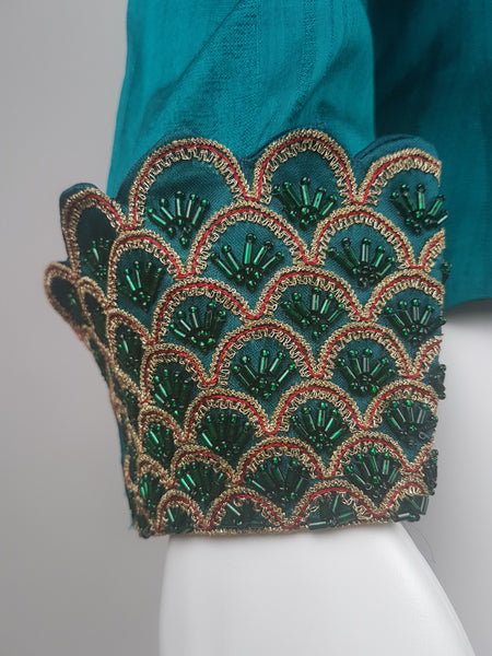Close-up of a deep teal silk cuff decorated with overlapping scallops, metallic-gold braid, red cord and emerald beads.