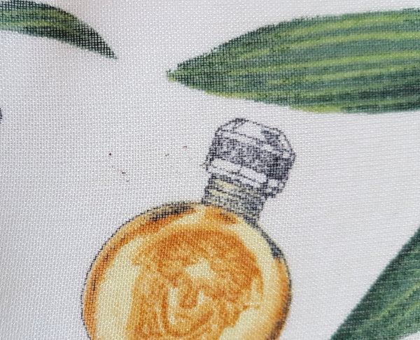 Close-up of green botanical branches and gold Versace perfume bottles, including a round Medusa-head bottle, printed on ivory chiffon.