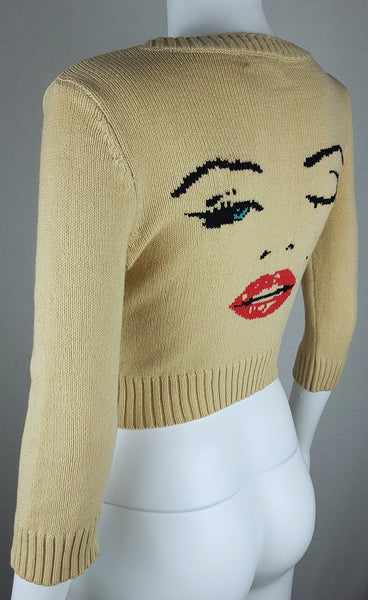 Three quarter back view of a vintage 2000s sand-beige Betsey Johnson cropped cardigan with three-quarter sleeves, cropped ribbed hem and with an intarsia Marilyn Monroe wink portrait in black, blue, red and white displayed on a mannequin