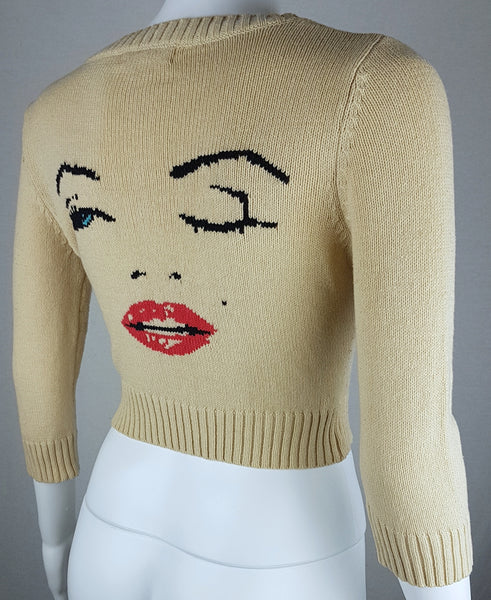 Three quarter back view of a vintage 2000s sand-beige Betsey Johnson cropped cardigan with three-quarter sleeves, cropped ribbed hem and with an intarsia Marilyn Monroe wink portrait in black, blue, red and white displayed on a mannequin