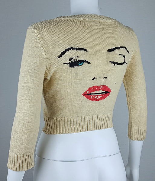 Three-quarter back view of a vintage 2000s sand-beige Betsey Johnson cropped cardigan with three-quarter sleeves, cropped ribbed hem and with an intarsia Marilyn Monroe wink portrait in black, blue, red and white displayed on a mannequin