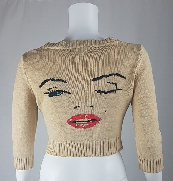 Full back view of a vintage 2000s sand-beige Betsey Johnson cropped cardigan with three-quarter sleeves, cropped ribbed hem and with an intarsia Marilyn Monroe wink portrait in black, blue, red and white displayed on a mannequin