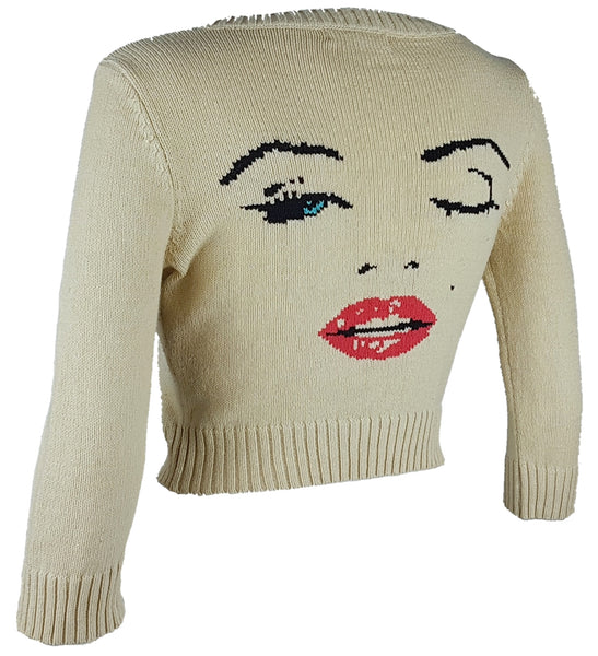 Three-quarter back view of a vintage 2000s sand-beige Betsey Johnson cropped cardigan with three-quarter sleeves, cropped ribbed hem and with an intarsia Marilyn Monroe wink portrait in black, blue, red and white displayed on a mannequin