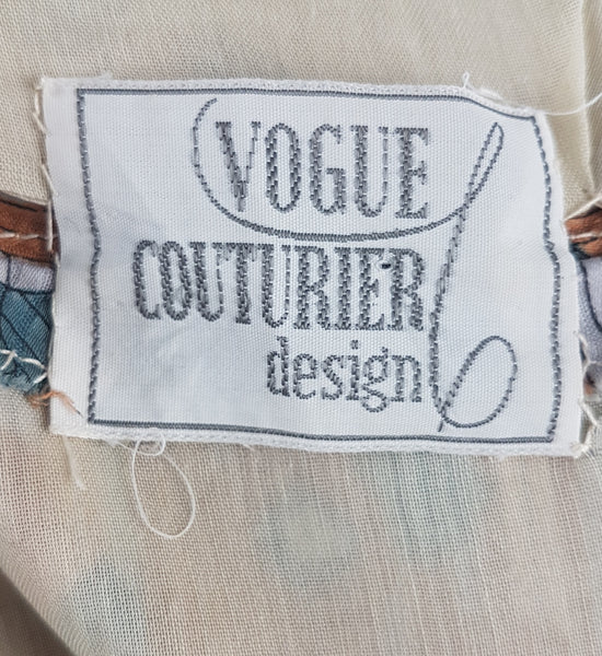 label detail view of a vintage late 1950s dressmaker-made Vogue Couturier Design tunic top in a tan and blue abstract floral print, with circular cape sleeves and a fitted waist displayed on a mannequin 
