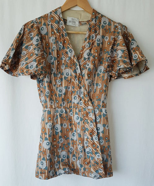 front view of a vintage late 1950s dressmaker-made Vogue Couturier Design tunic top in a tan and blue abstract floral print, with circular cape sleeves and a fitted waist displayed on a hanger to show true neckline 