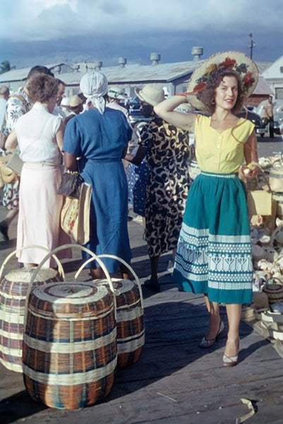 "American in Cuba, 1950s by Heinrich Heidersberger." Vintage colour photograph of a woman in a yellow blouse and teal patterned skirt carrying a large woven basket through a busy outdoor market.