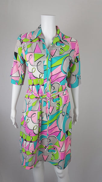 Front view of Vintage 1980s Lady Bayard shirt dress with short sleeves, a pointed collar, button front, matching tie belt, and a bold abstract geometric print in pink, turquoise, lime green, grey, and black displayed on a mannequin