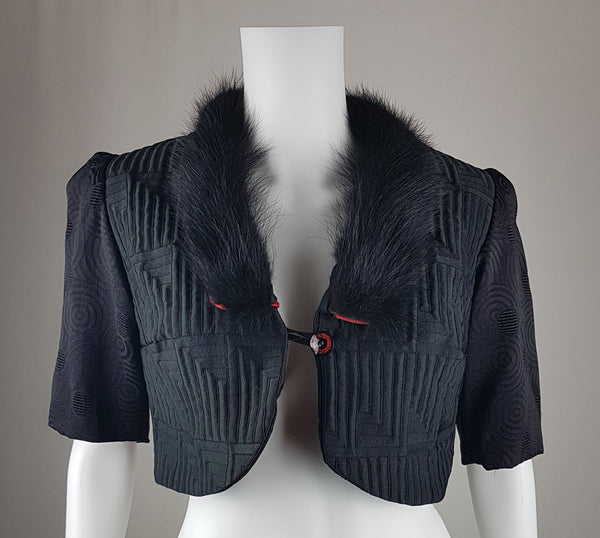 Vintage black brocade cropped bolero jacket with a fur collar, red satin lining, ribbon loop and decorative button displayed on a mannequin - front view